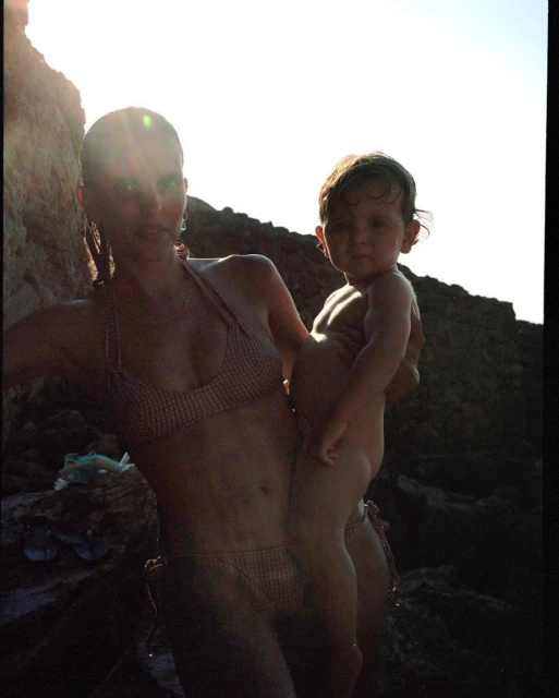 Woman in a bikini holding a baby on a rocky beach with sunlight, creating a sunlit portrait in a natural outdoor environment.