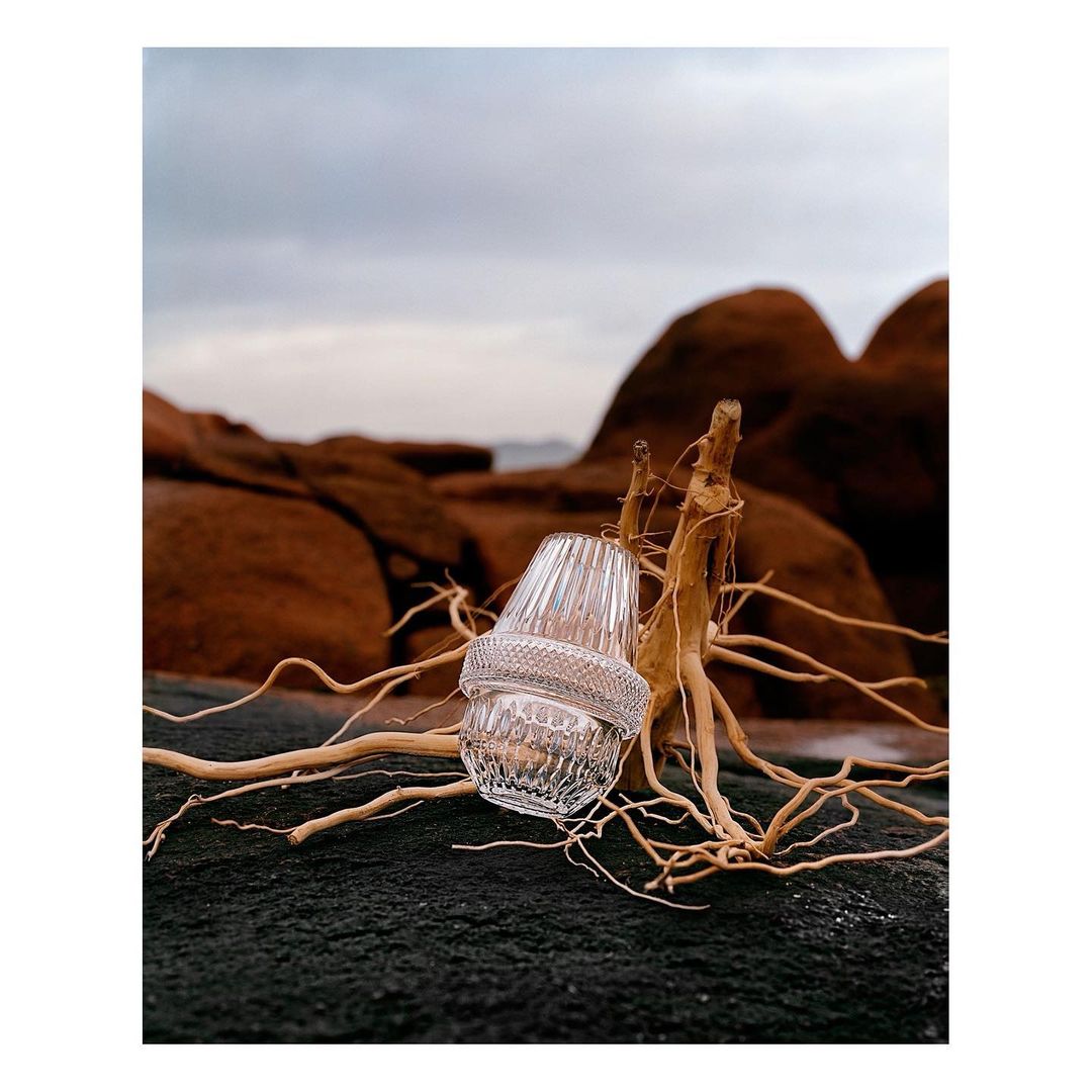 Clear glass candle holders sit on a dark rock near weathered driftwood, backed by tan rocks and a cloudy sky, creating a serene scene.