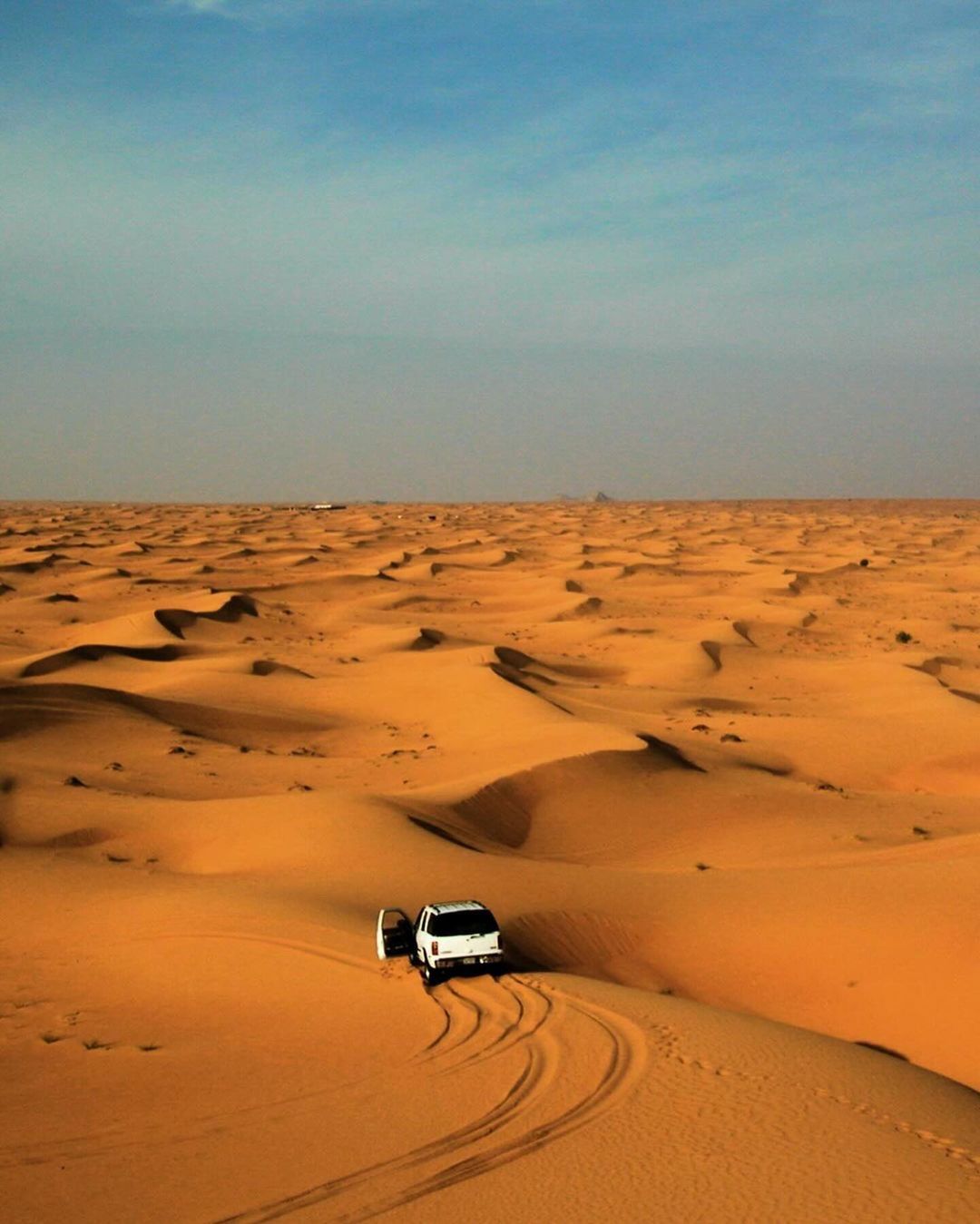 Off-road vehicle drives across the undulating sand dunes of a desert landscape beneath a clear blue sky, tire tracks visible.
