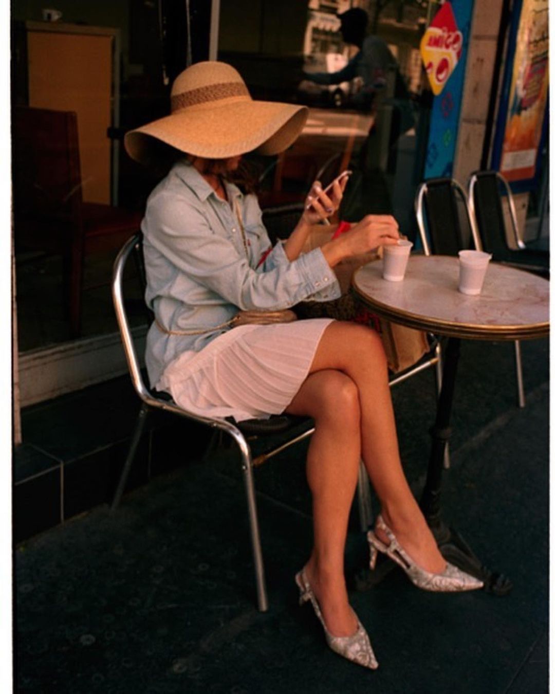 Woman in a wide-brimmed hat sits at a table using her phone, with two cups on the table, in an outdoor setting.