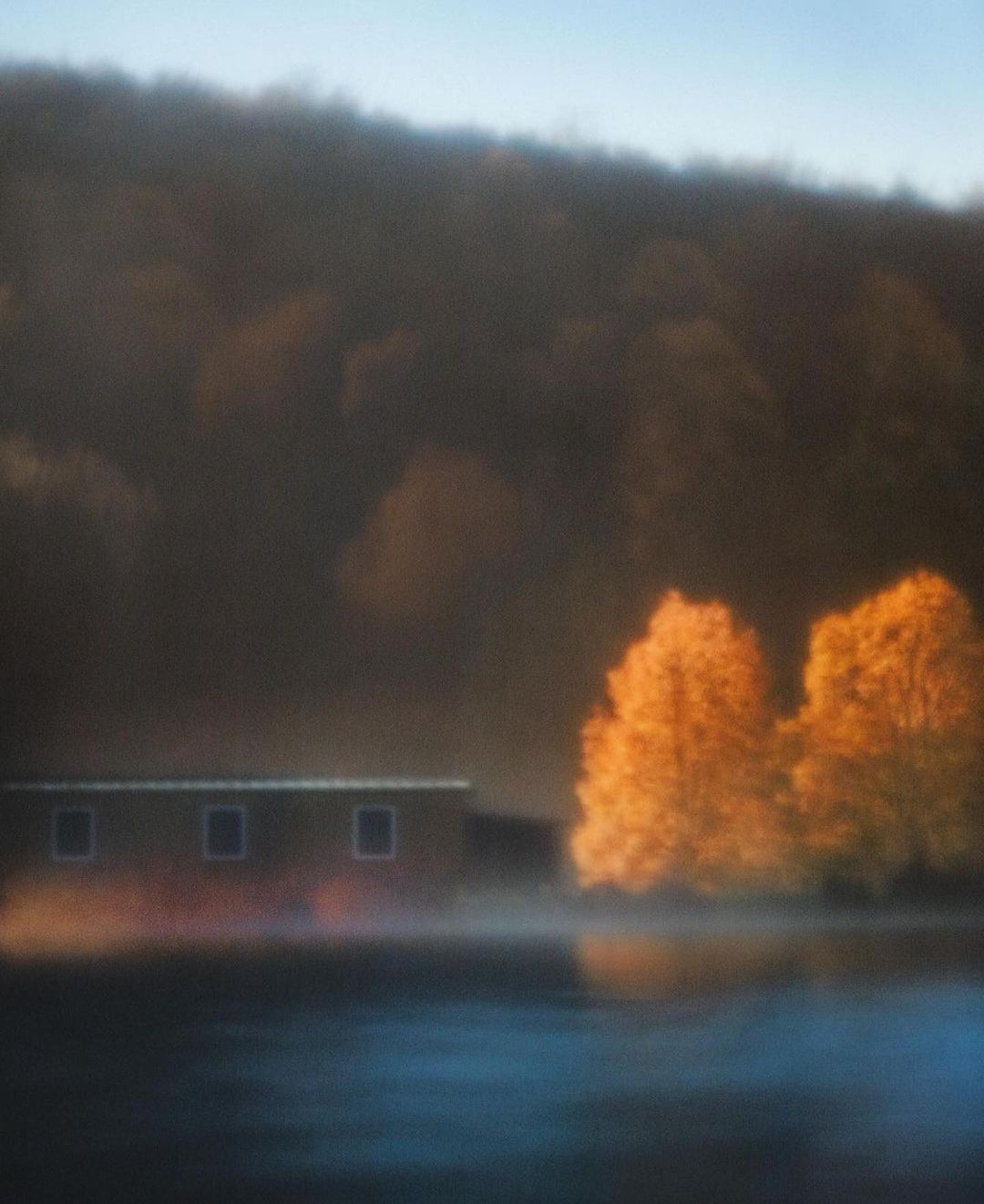 Scenic landscape featuring a tranquil lake reflecting golden autumn trees, with a building nestled against a hillside in soft, misty light.