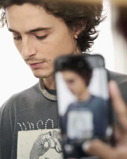 Portrait of a young adult male with wavy hair wearing a gray graphic tee, partially obscured by a hand holding a smartphone.