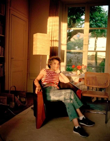 Mature woman in striped shirt, jeans, and sneakers sits in an armchair in a sunlit room with a window, books, and a wooden chair.