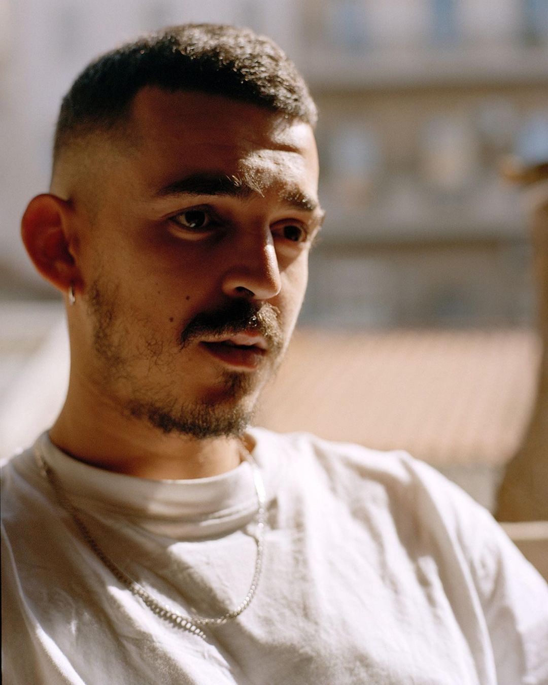 Close-up portrait of a young adult male with a buzzcut, wearing a white t-shirt and silver chain necklace, posing outdoors in natural light.