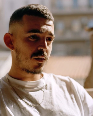 Close-up portrait of a young adult male with a buzzcut, wearing a white t-shirt and silver chain necklace, posing outdoors in natural light.