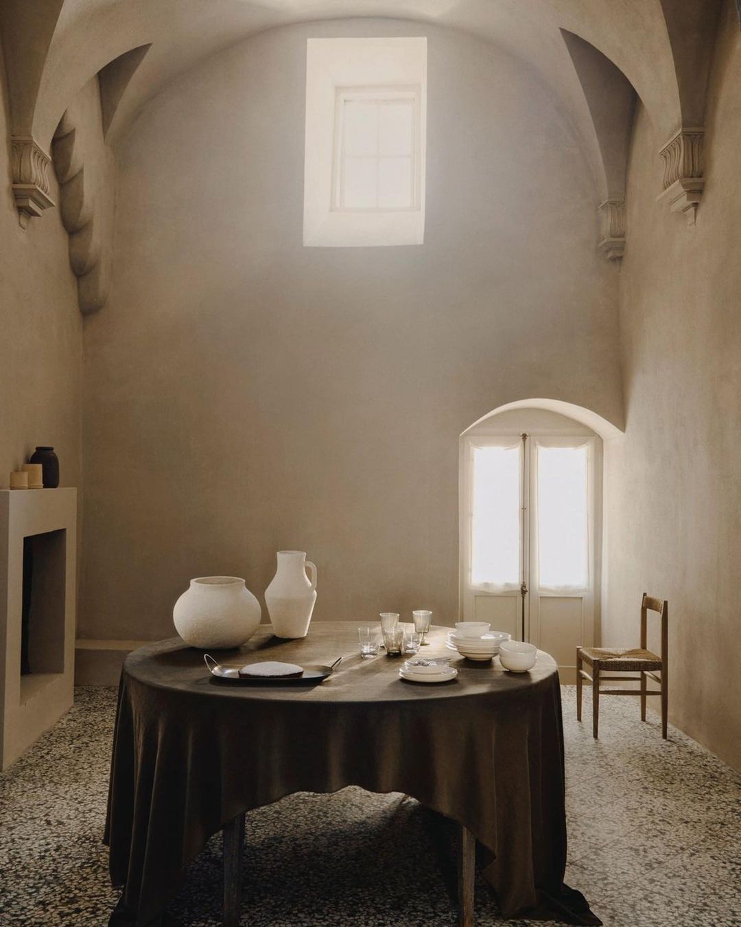 Interior scene featuring a draped round table set with plates and glasses in a room with textured walls, arched ceilings, and natural light.