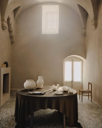 Interior scene featuring a draped round table set with plates and glasses in a room with textured walls, arched ceilings, and natural light.