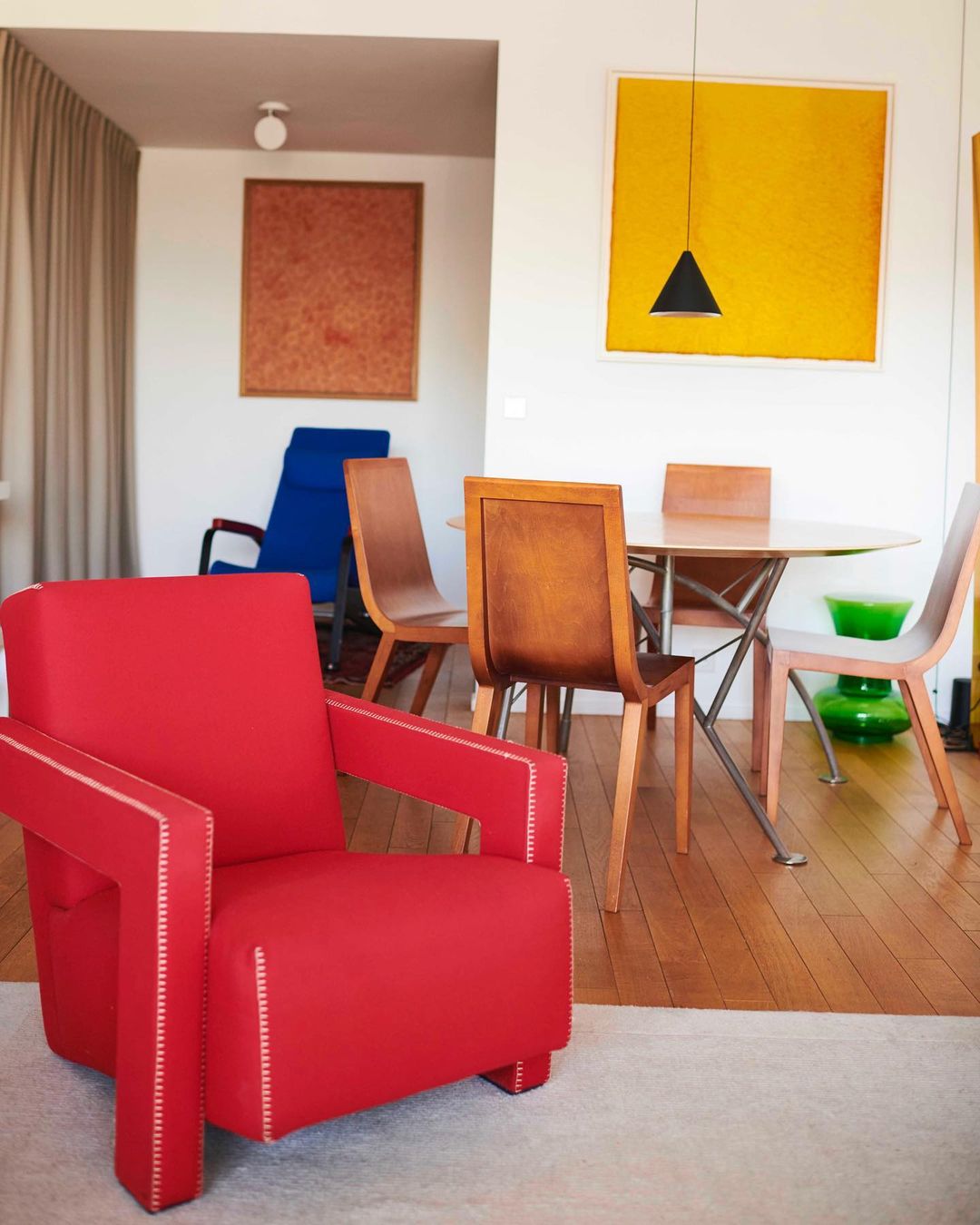 Bright interior with a red modern chair, a wood table surrounded by chairs, and abstract paintings in yellow and brown hues.