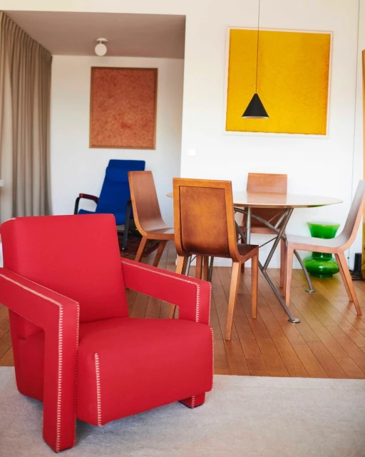 Bright interior with a red modern chair, a wood table surrounded by chairs, and abstract paintings in yellow and brown hues.