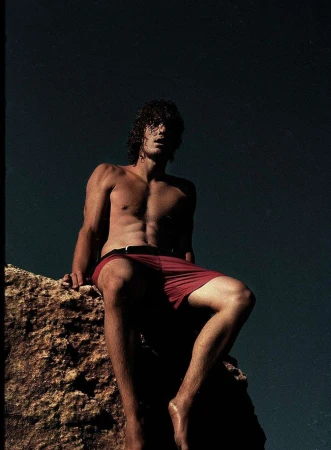 Shirtless man with curly hair wearing red swim shorts seated on a rocky outcrop under a dark sky.