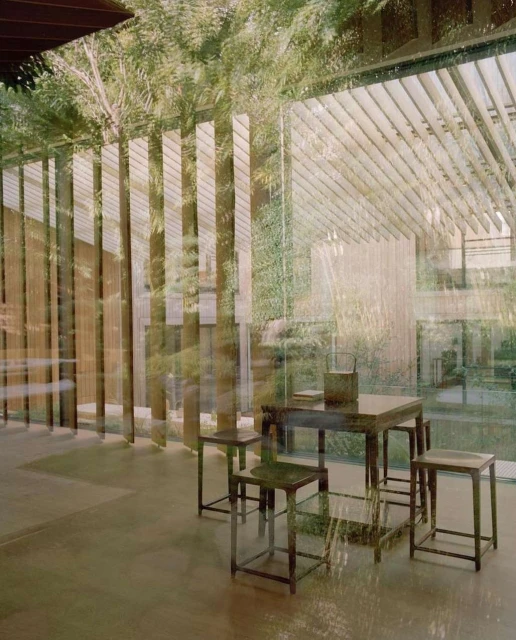 Indoor scene featuring a dark wooden table and four stools. The interior space has an open feel, with a window view of outdoor foliage.