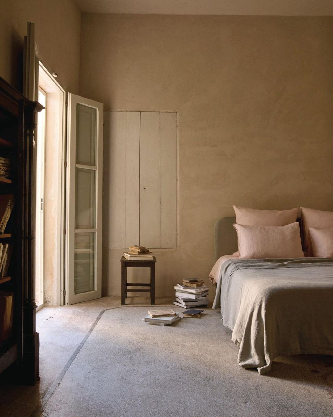 A sunlit bedroom interior featuring a made bed with neutral linens, a stack of books on the floor, and an open balcony door.