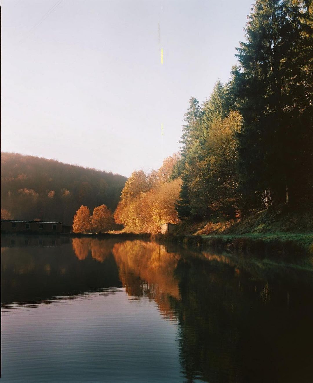 Still lake reflects autumnal trees and building on a sunny day. Evergreen trees stand on the right. Overall soft, desaturated tones.