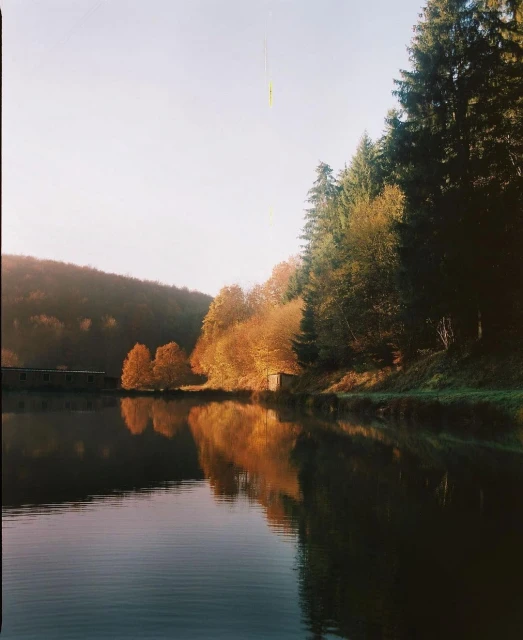 Still lake reflects autumnal trees and building on a sunny day. Evergreen trees stand on the right. Overall soft, desaturated tones.