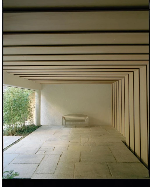 Minimalist interior architecture featuring a silver bench, stone flooring, and a modern slatted ceiling design in a sunlit space.