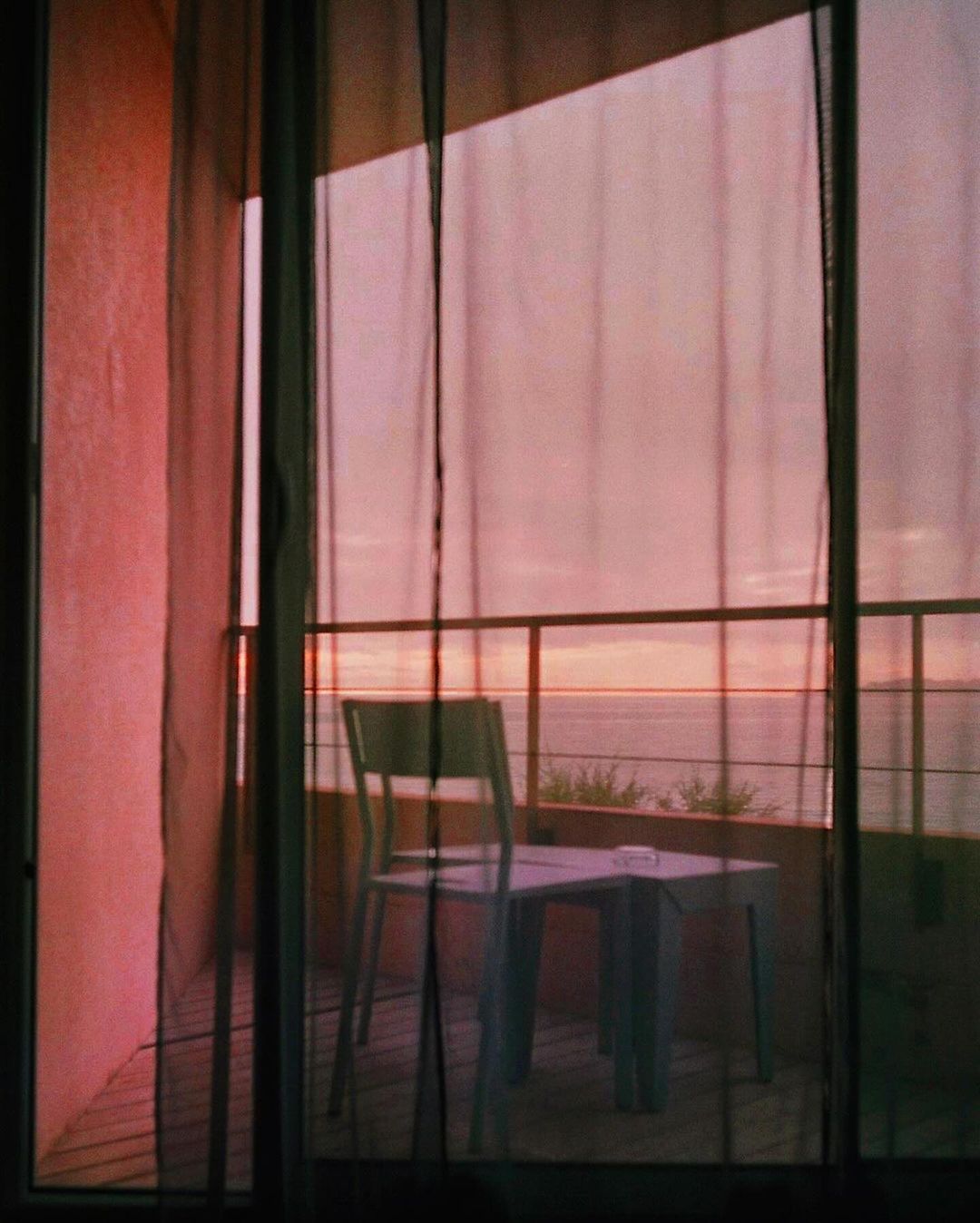 Chair and table on balcony with black netting overlooking ocean at sunset with soft, pink light and pastel color palette.
