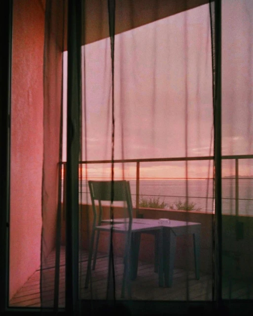 Chair and table on balcony with black netting overlooking ocean at sunset with soft, pink light and pastel color palette.