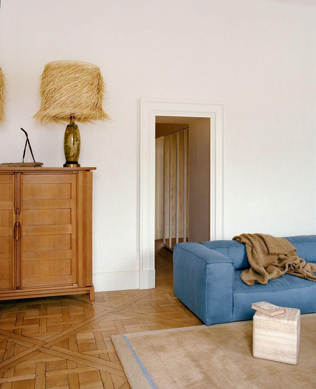 Living room interior featuring a blue sofa, light brown rug, a wooden cabinet with a lamp, and a doorway in a minimal aesthetic.