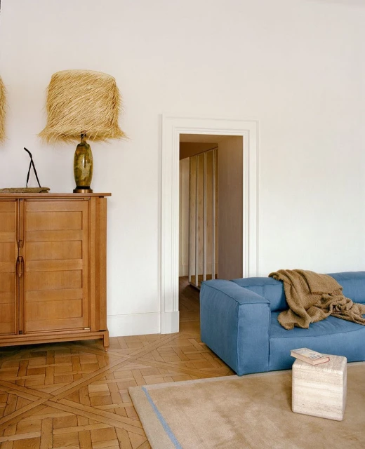 Living room interior featuring a blue sofa, light brown rug, a wooden cabinet with a lamp, and a doorway in a minimal aesthetic.