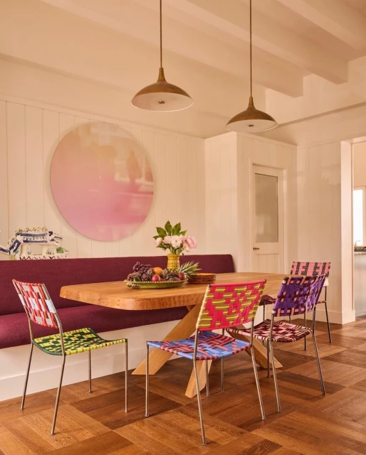 Dining area featuring a wooden table with fruit, colorful chairs, overhead lighting, and pink-toned wall art in a bright, modern interior.