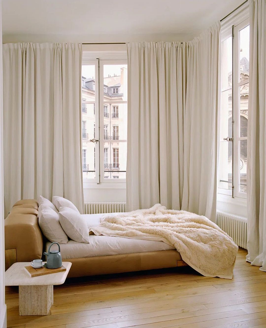 Sunlit bedroom interior with a tan platform bed, white bedding, and a small travertine side table with a teapot, creating a cozy setting.