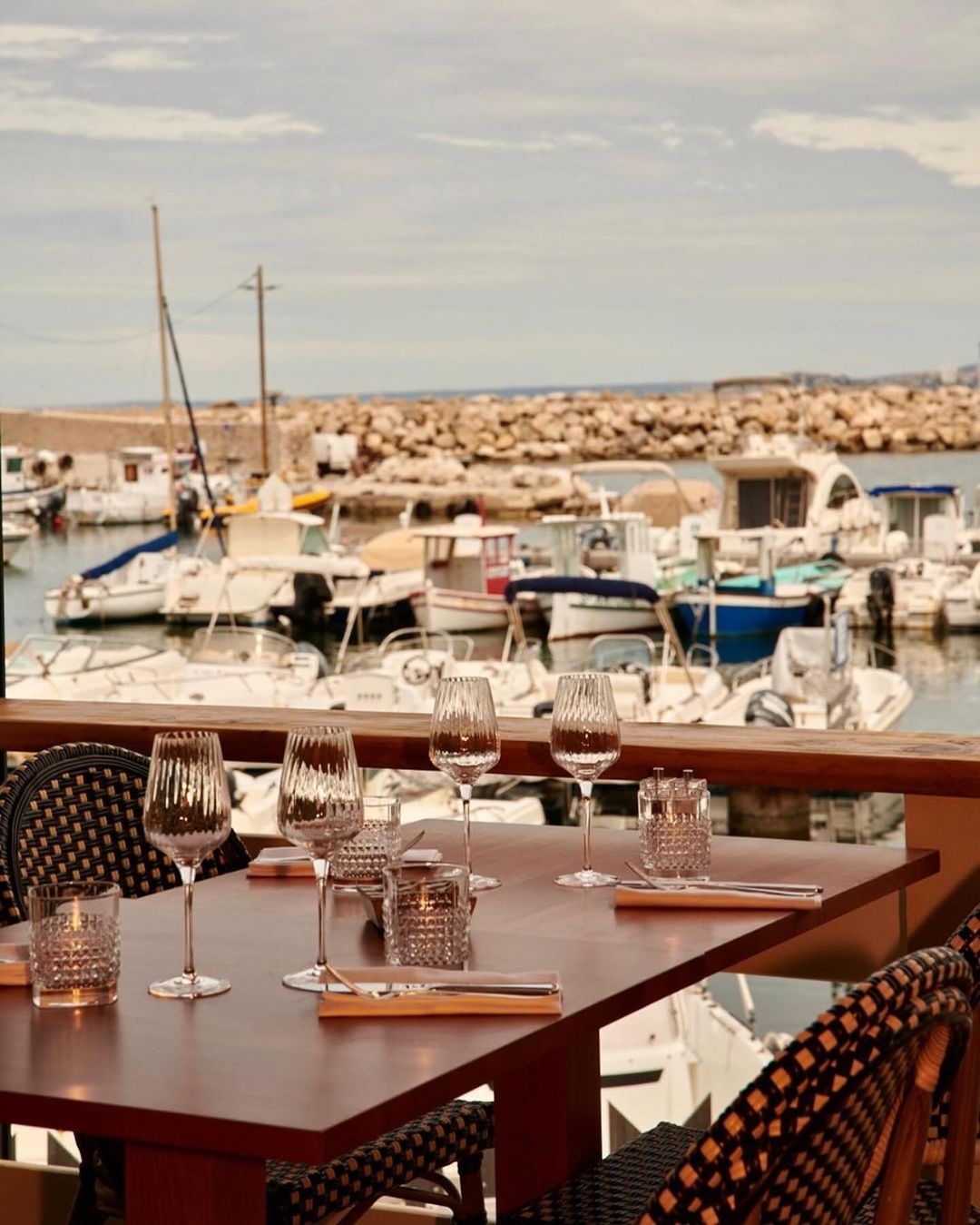 Outdoor restaurant table set for dining with crystal glasses, silverware, and harbor view with boats and a stone breakwater in the background.