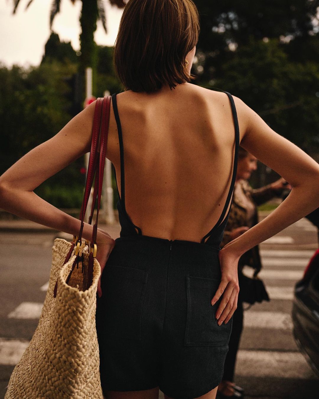 Back view of a young woman with short hair wearing a black jumpsuit, holding a straw bag, in an urban outdoor environment.