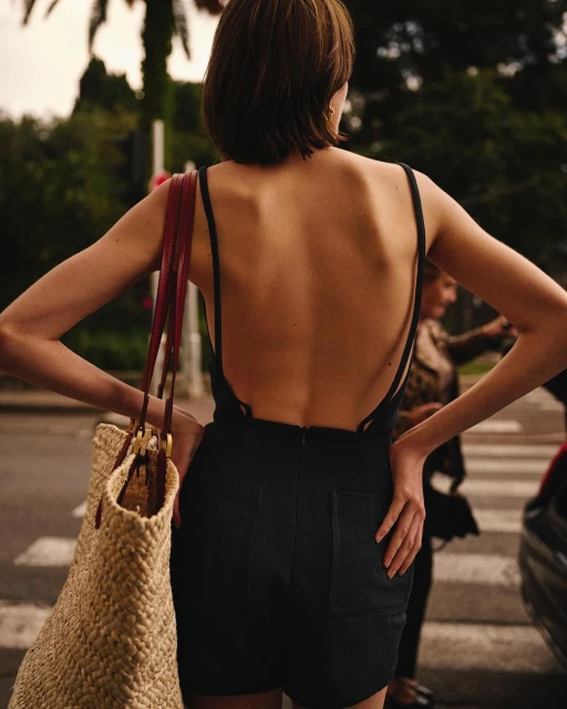 Back view of a young woman with short hair wearing a black jumpsuit, holding a straw bag, in an urban outdoor environment.