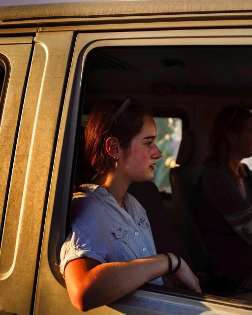 Woman gazes out of a car window with warm sunlight casting over the vehicle's exterior and interior, showcasing a candid moment.