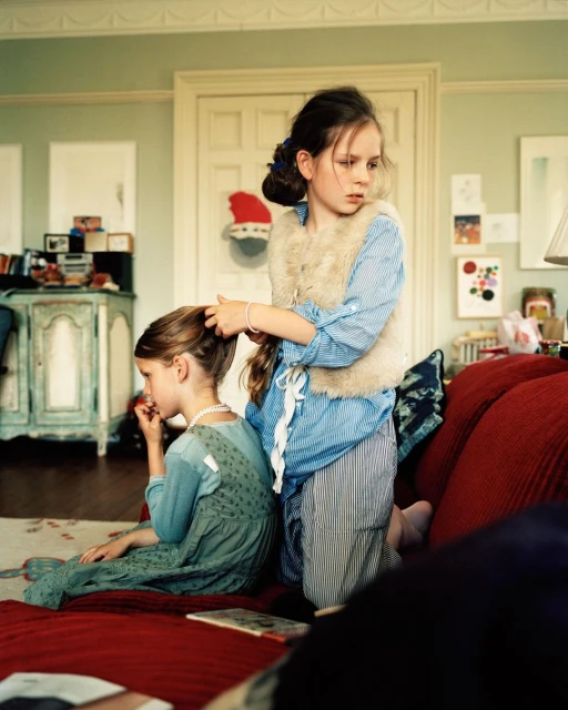 Two young girls in a home interior, one is seated in a teal dress while the other stands in a blue striped shirt and braids her hair.