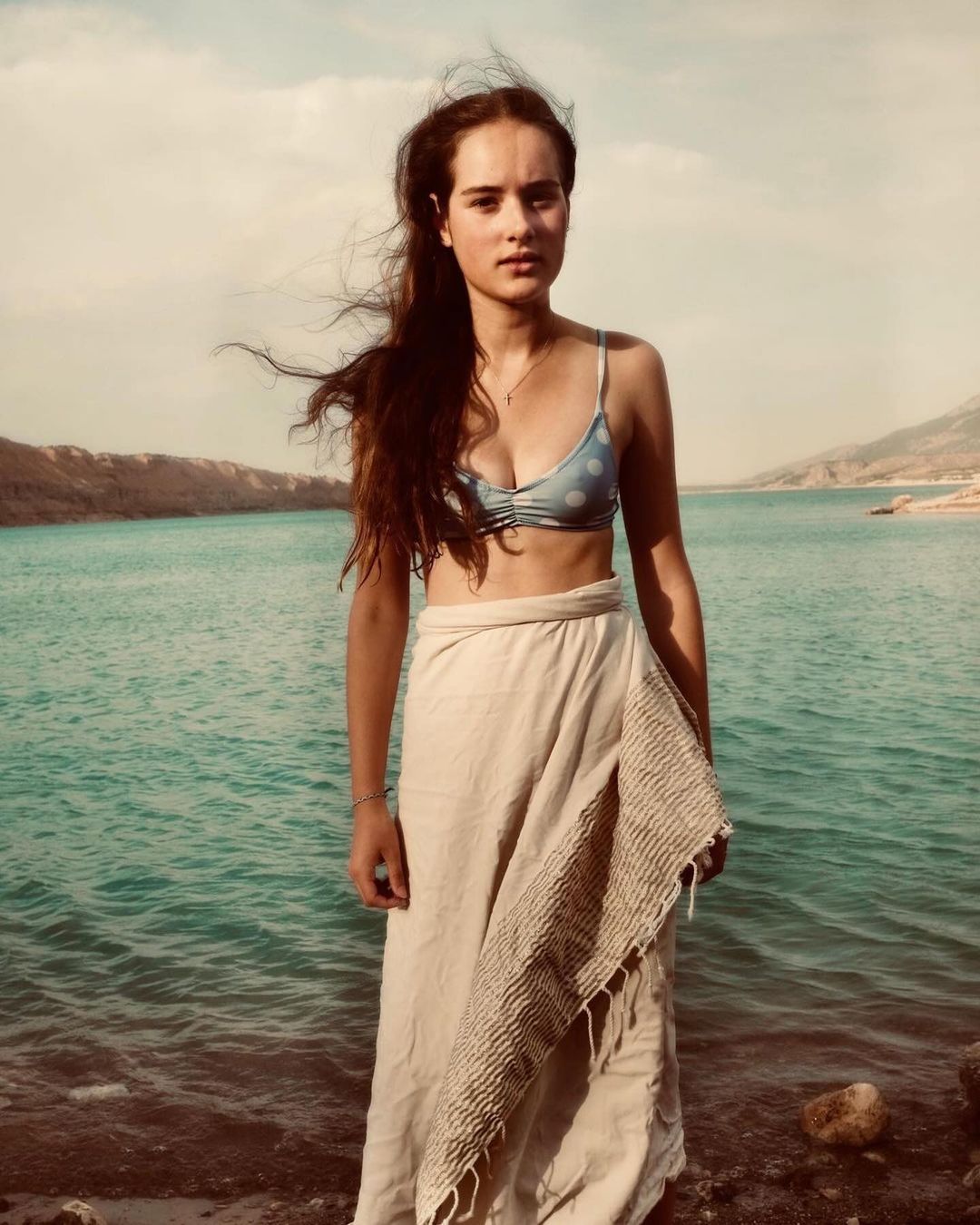 Young woman stands on a rocky beach with turquoise water and mountains in the background, wearing a sarong and blue polka dot bikini top.