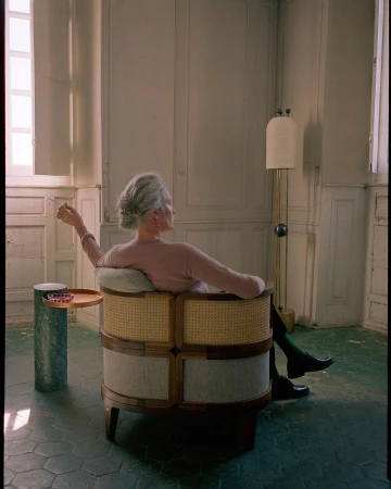 An elderly woman with grey hair seated in a round cane armchair holding a cigarette inside a room with panelled walls.