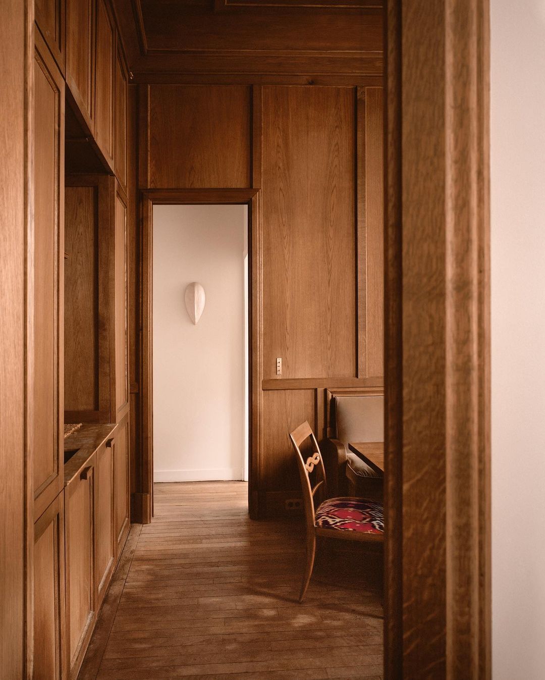 Interior design of a wood-paneled room featuring a vintage-style chair and a glimpse into a white-walled area with a wall sculpture.