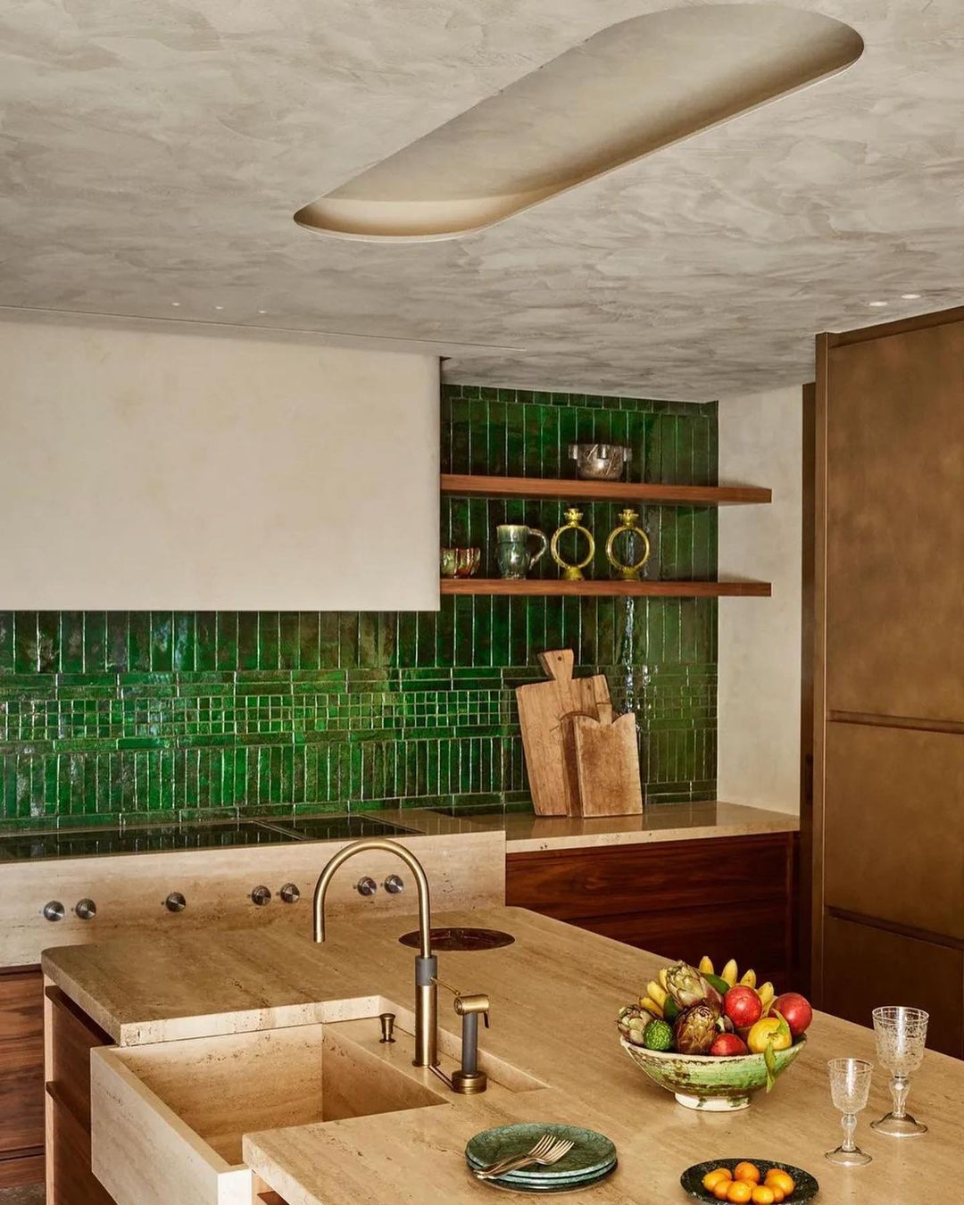 A stylish kitchen interior features green backsplash tiles, a beige countertop island with a sink, and fruit in a decorative bowl.