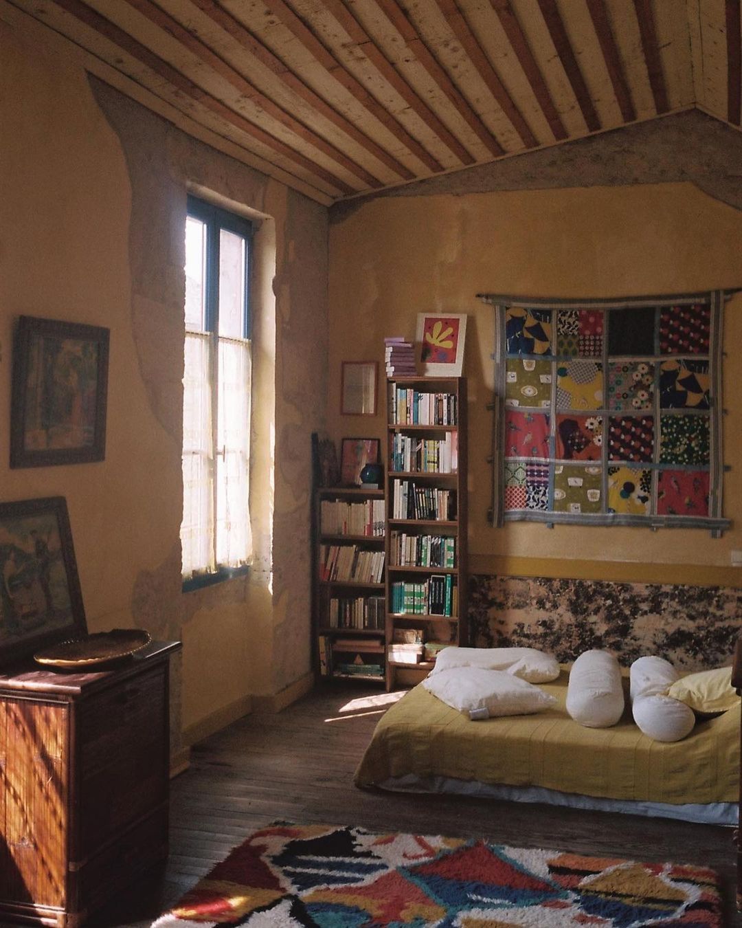 Sunlit bedroom interior with a bed, bookcase filled with books, and a vibrant, geometrically patterned rug on the wooden floor.