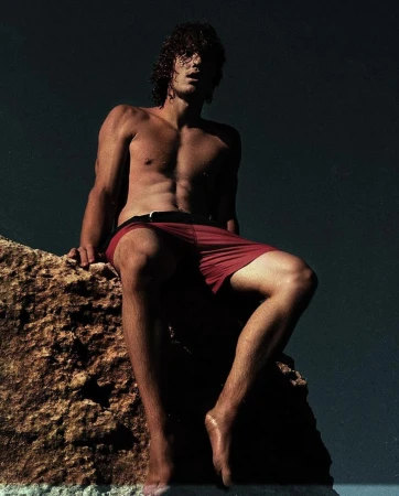 Athletic male model sits shirtless on a textured rock with a dark teal background in swimwear fashion, portrait, lifestyle, beach style.