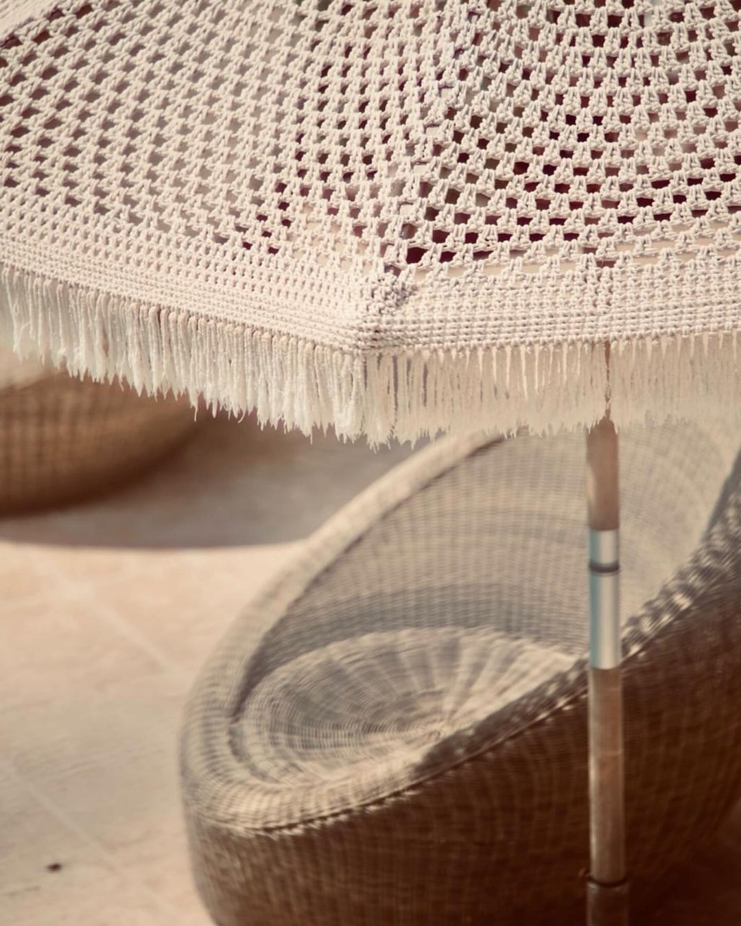 Creamy white macrame sun umbrella casts shadows on a woven chair on a neutral toned patio on a sunny day. Vacation vibes.