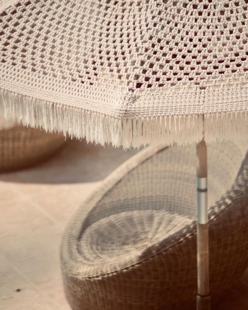 Creamy white macrame sun umbrella casts shadows on a woven chair on a neutral toned patio on a sunny day. Vacation vibes.
