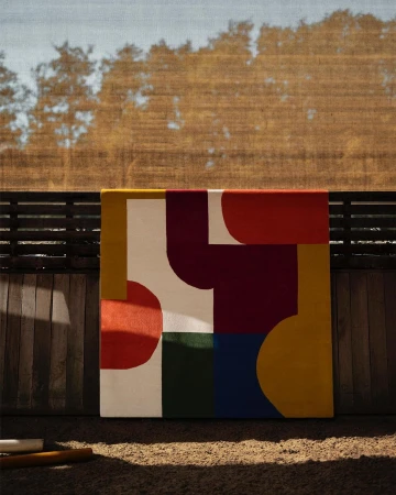 Abstract painting with red, white, and yellow geometric shapes, positioned against a fence with trees and a blue sky in the background.