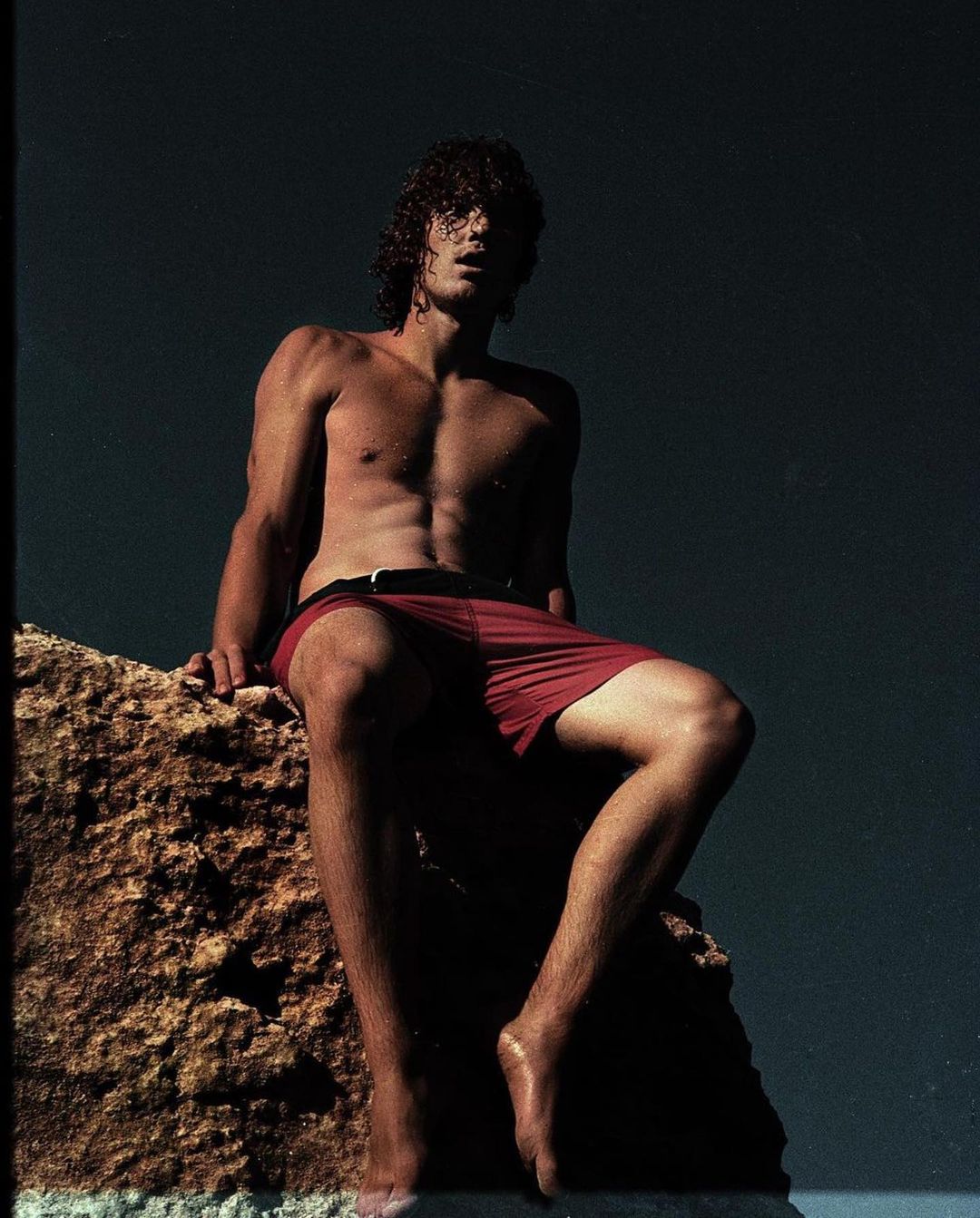 Man sitting shirtless on a rock in red swim trunks. Muscular physique, sunlit skin, dark curly hair, against a dark background.