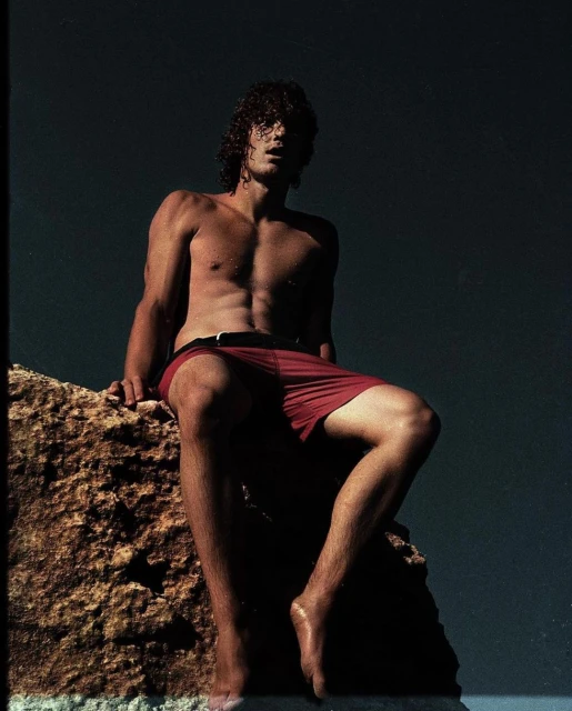 Man sitting shirtless on a rock in red swim trunks.  Muscular physique, sunlit skin, dark curly hair, against a dark background. 