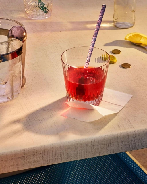 Red cocktail in a clear glass with a patterned straw on a white napkin next to a lemon peel and bar accessories.
