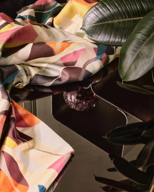 Still life composition: patterned textile, glossy leaves, and a faceted glass object on a dark reflective surface, indoor setting.