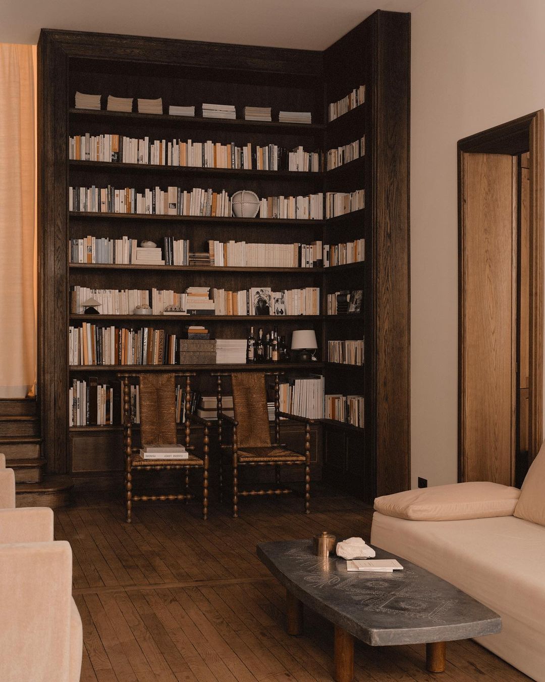 Interior view of a living room featuring a large bookcase filled with books, two woven chairs, a stone coffee table, and a cream-colored sofa.
