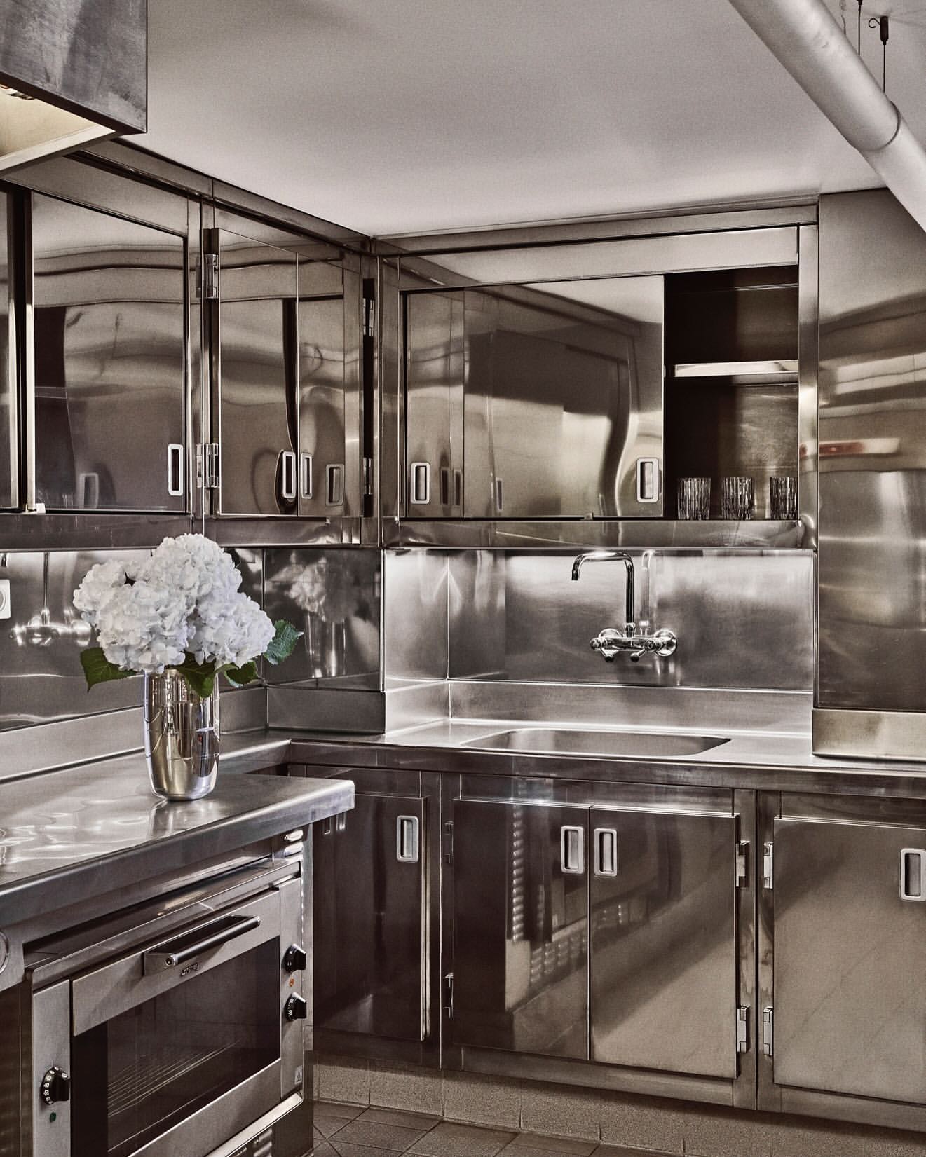 Stainless steel kitchen with cabinets, drawers, sink, faucet, oven, tiled floor, and silver vase of white flowers.