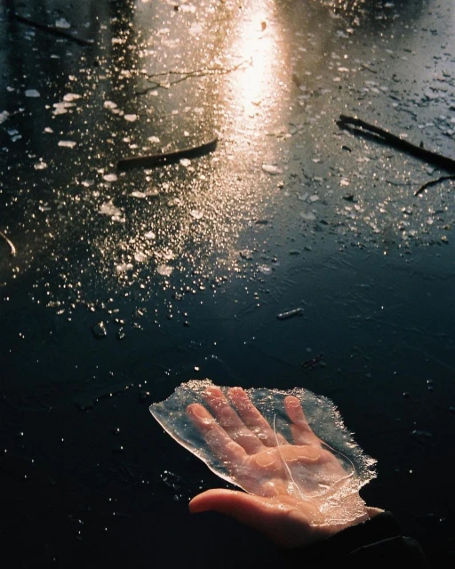 Close-up of a human hand holding a piece of transparent ice against a dark, frozen, and reflective background with patches of sunlight.