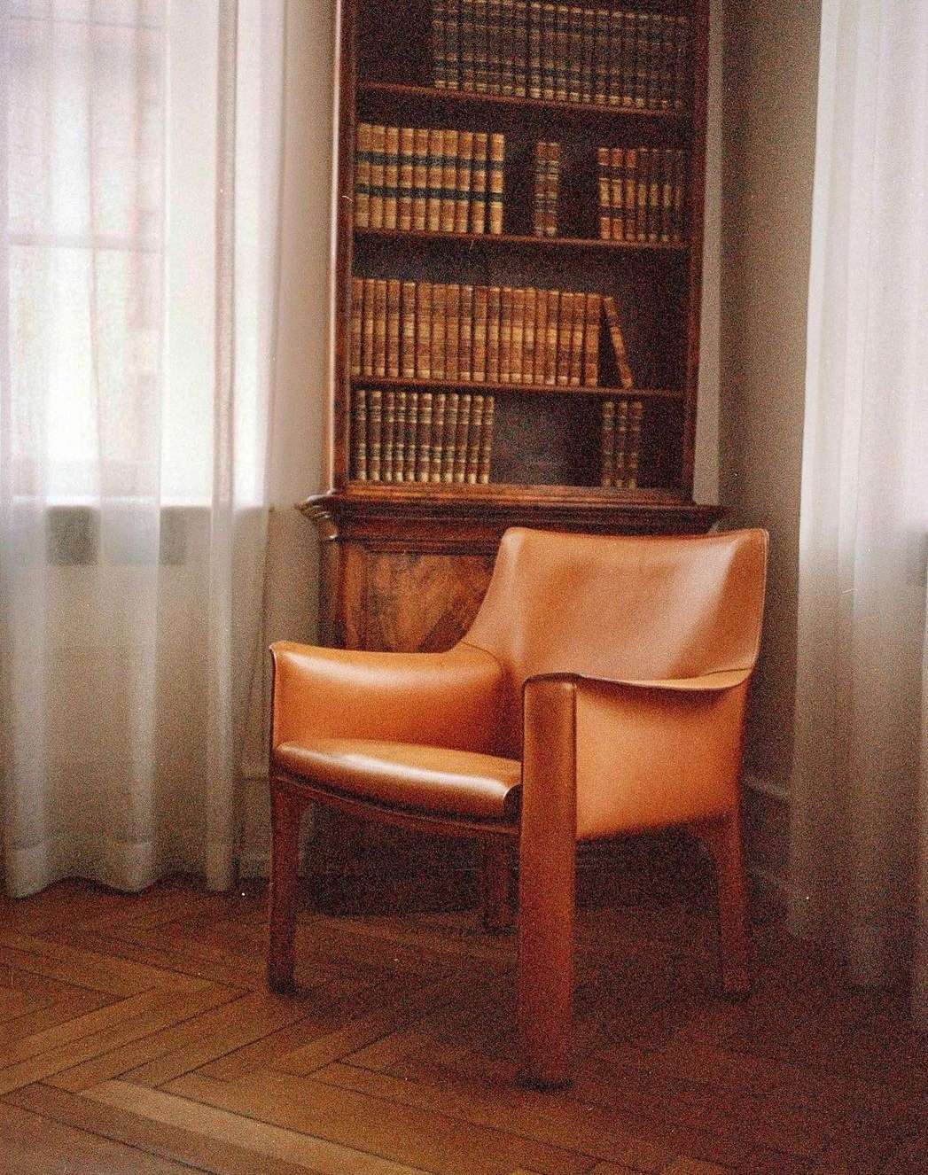Interior view of a tan leather chair on a wood floor, positioned near a wooden bookcase and a window with white curtains.