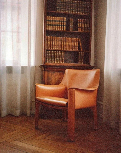 Interior view of a tan leather chair on a wood floor, positioned near a wooden bookcase and a window with white curtains.