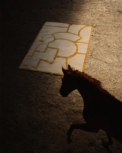 A brown horse stands on a tan surface, with a graphic rug behind it, lit by soft light that creates a visually appealing contrast.