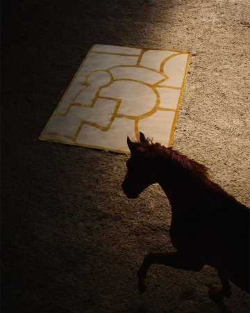 A brown horse stands on a tan surface, with a graphic rug behind it, lit by soft light that creates a visually appealing contrast.
